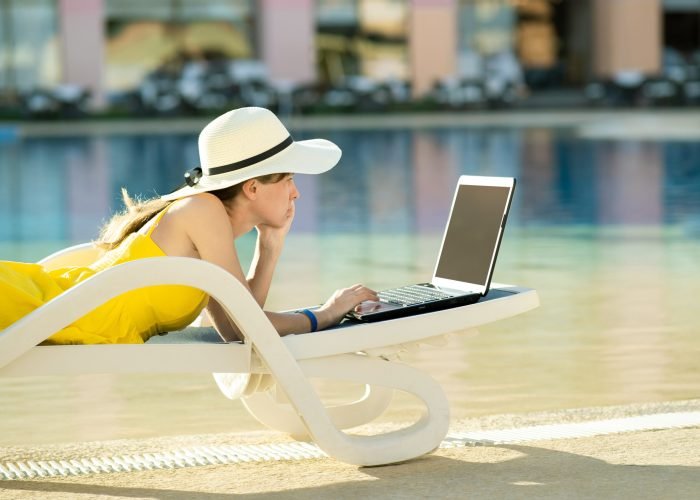 Young woman is lying on beach chair working on computer laptop connected to wireless internet typing text on keys in summer resort. Doing business while travelling concept.